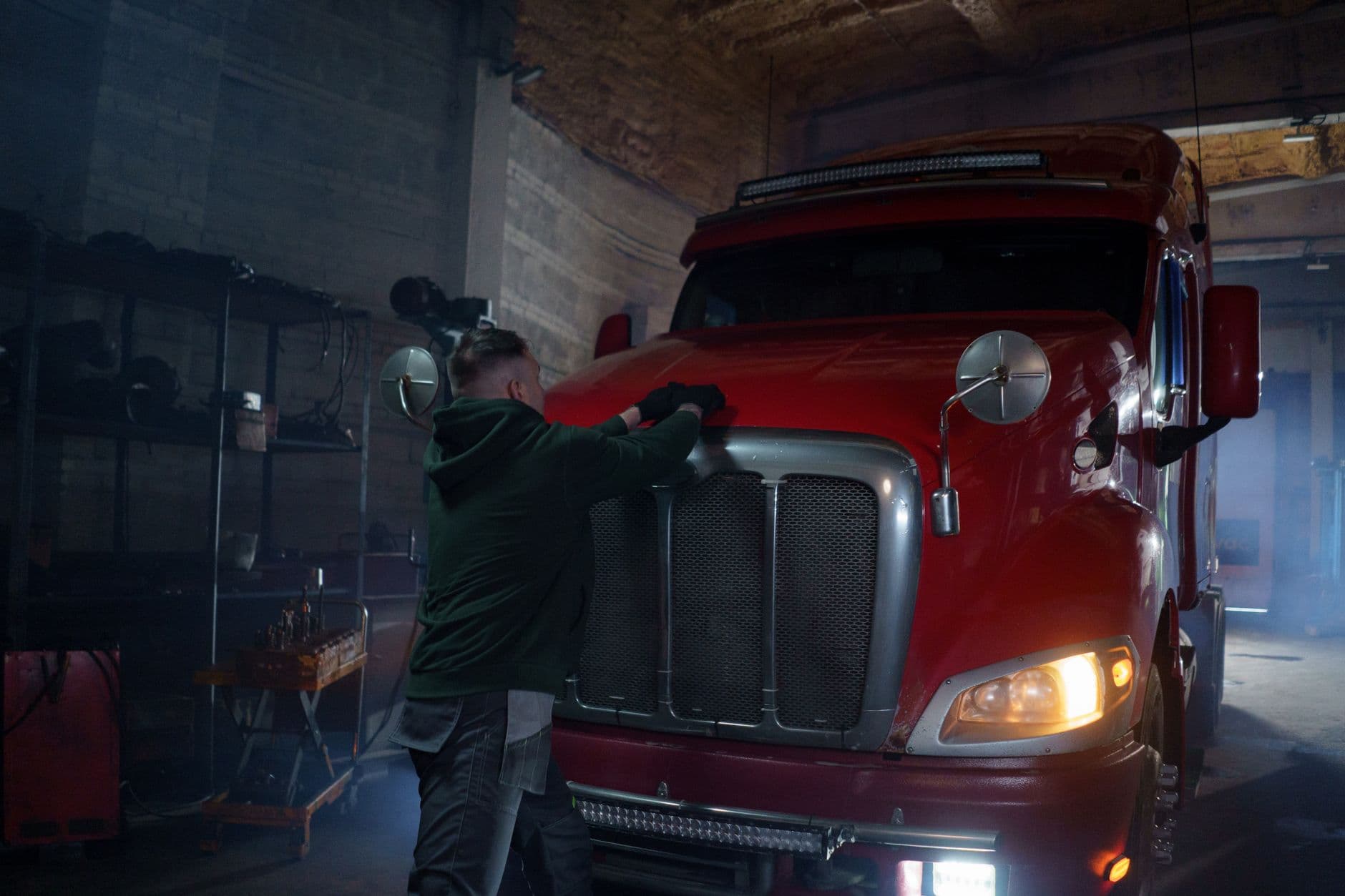 A mechanic in a dimly lit garage working on a red semi truck, focus on repair.